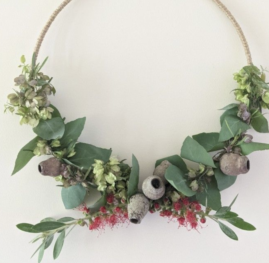Natural wreath with green foliage, gum nuts and red blossoms arranged on a circular hanging frame.